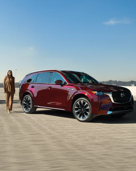 Mazda CX-90 SUV de lujo en color rojo estacionada sobre superficie desértica, con una mujer caminando junto al vehículo bajo un cielo azul despejado.
