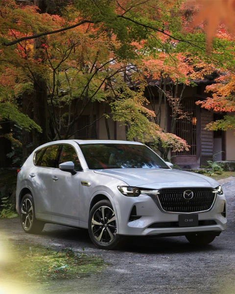 Mazda CX-60 híbrida enchufable color blanco perla estacionada en un entorno otoñal con diseño arquitectónico japonés al fondo.