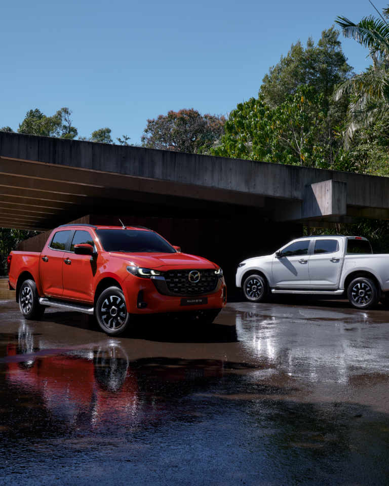 Dos Mazda BT-50 2025, una roja y una blanca, estacionadas bajo estructura moderna con fondo tropical.